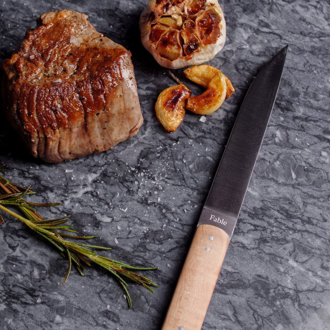 Steak, garlic, rosemary, and a knife on a dark surface